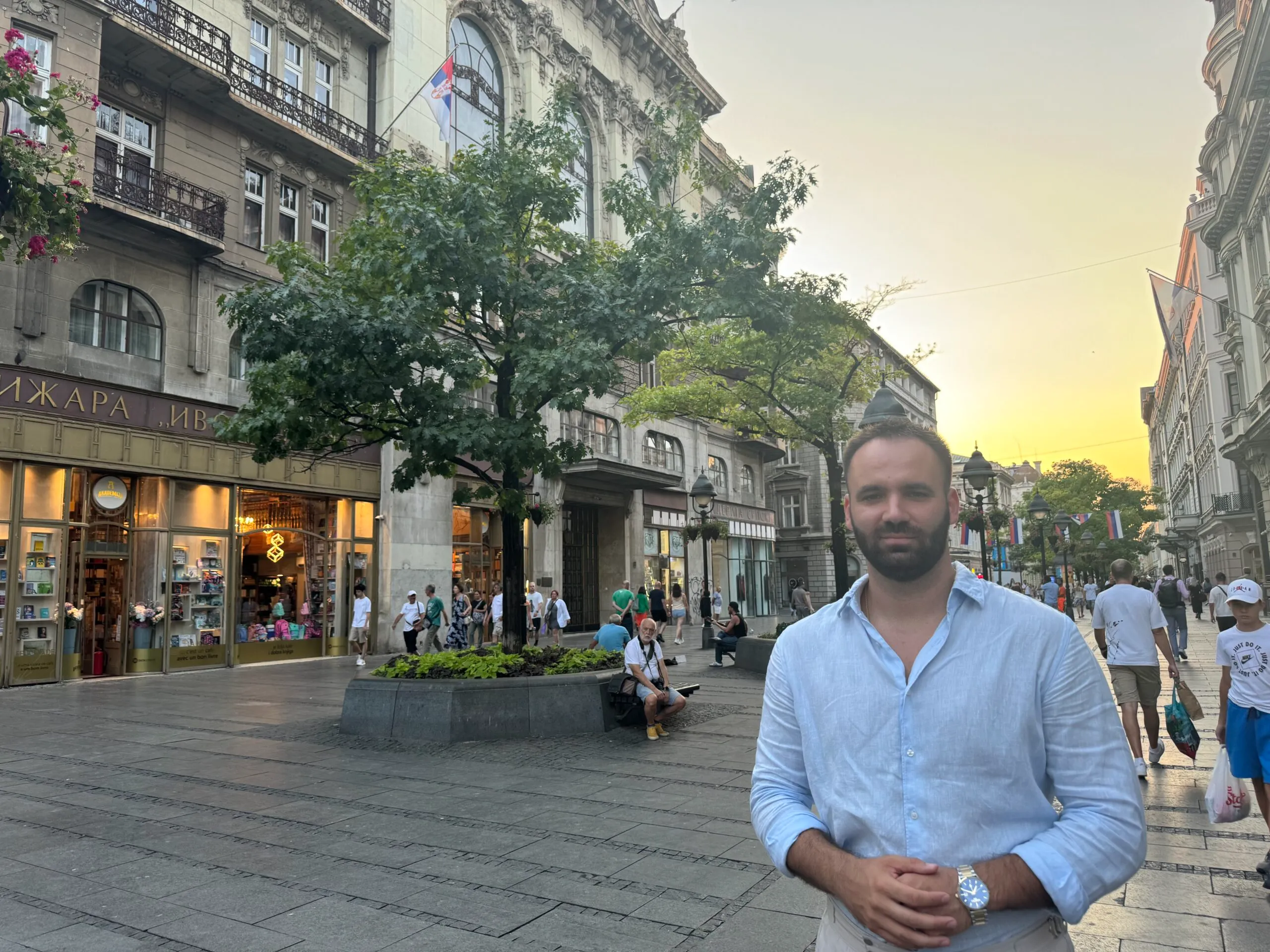 Centar Beograda sa poznatom knjižarom i prolaznicima u sumrak, muškarac u plavoj košulji u prvom planu.